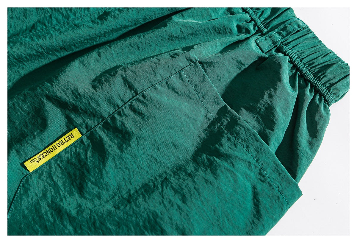 Close-up of teal-green trousers showing a large pocket, gathered elastic waistband, and small yellow woven label.