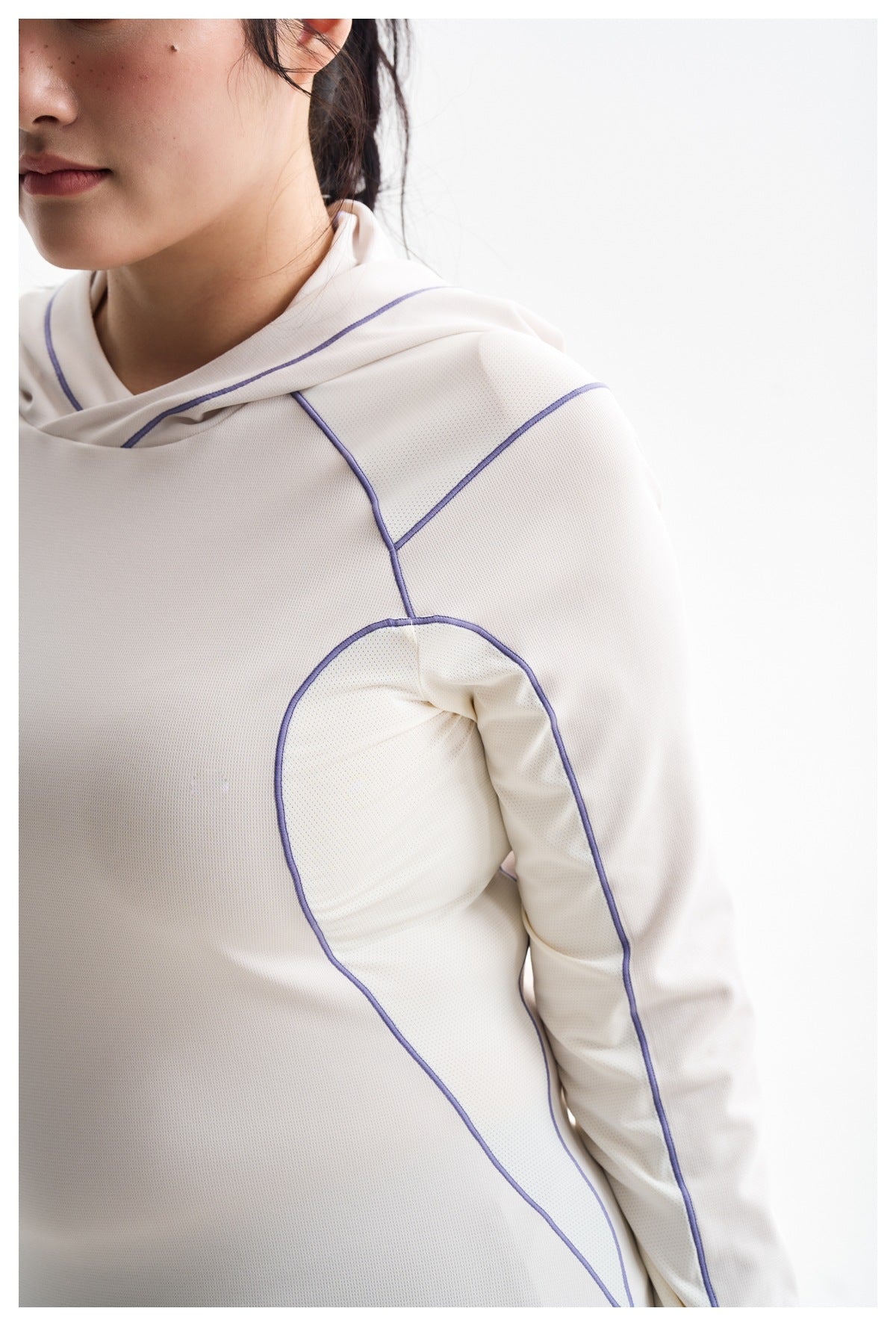 Close-up of an ivory hooded top showing its fine perforated texture and purple piping at the shoulder and underarm.
