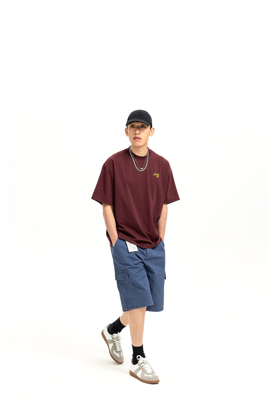 Full-length model wearing a burgundy oversized T-shirt with small yellow chest script and blue cargo shorts.