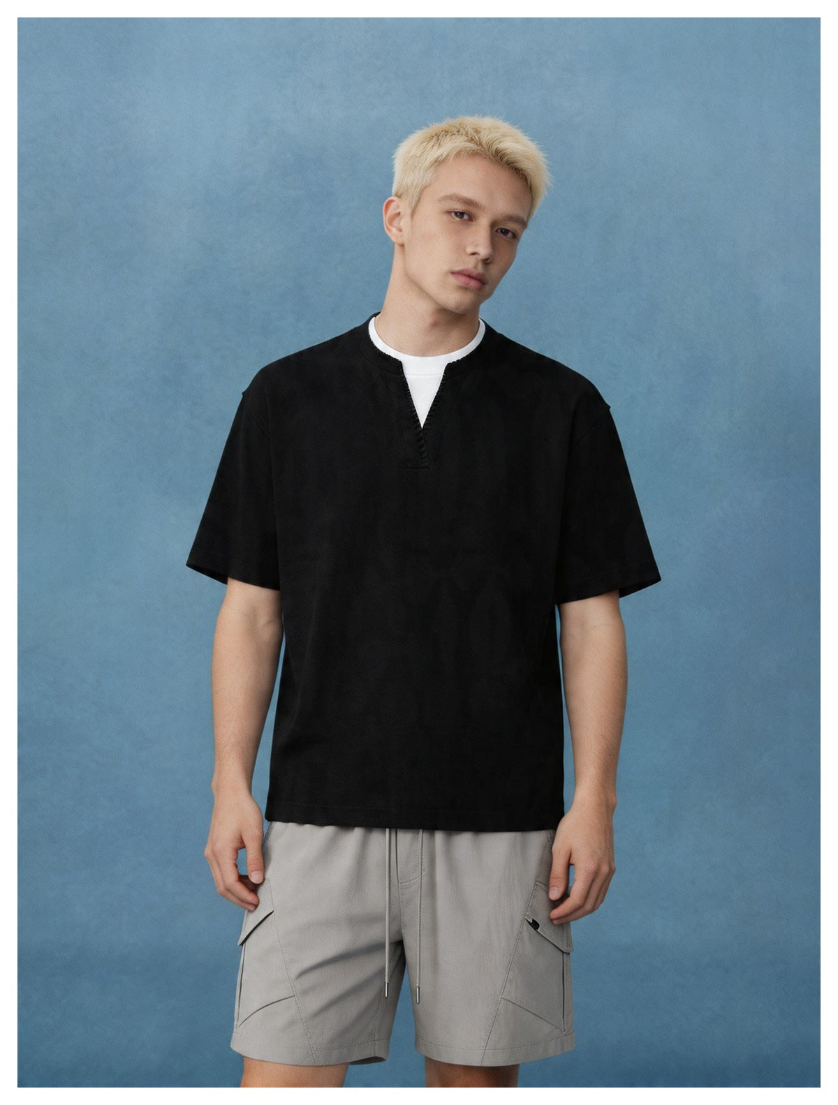 Man wearing a black notched V-neck T-shirt with white under-layer and gray cargo shorts against a blue backdrop