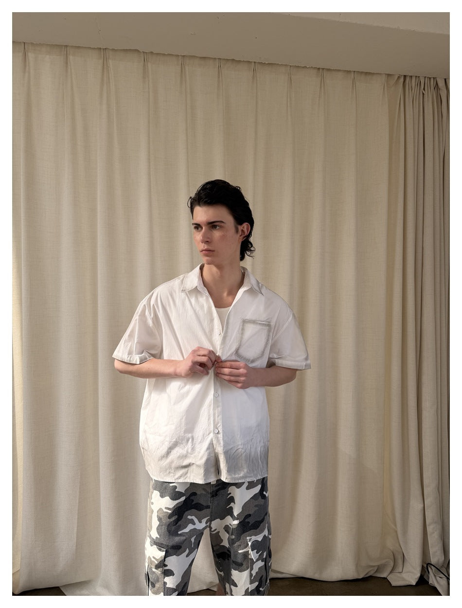 Model fastening a white short-sleeve button-up shirt with a chest pocket, paired with gray camouflage shorts.