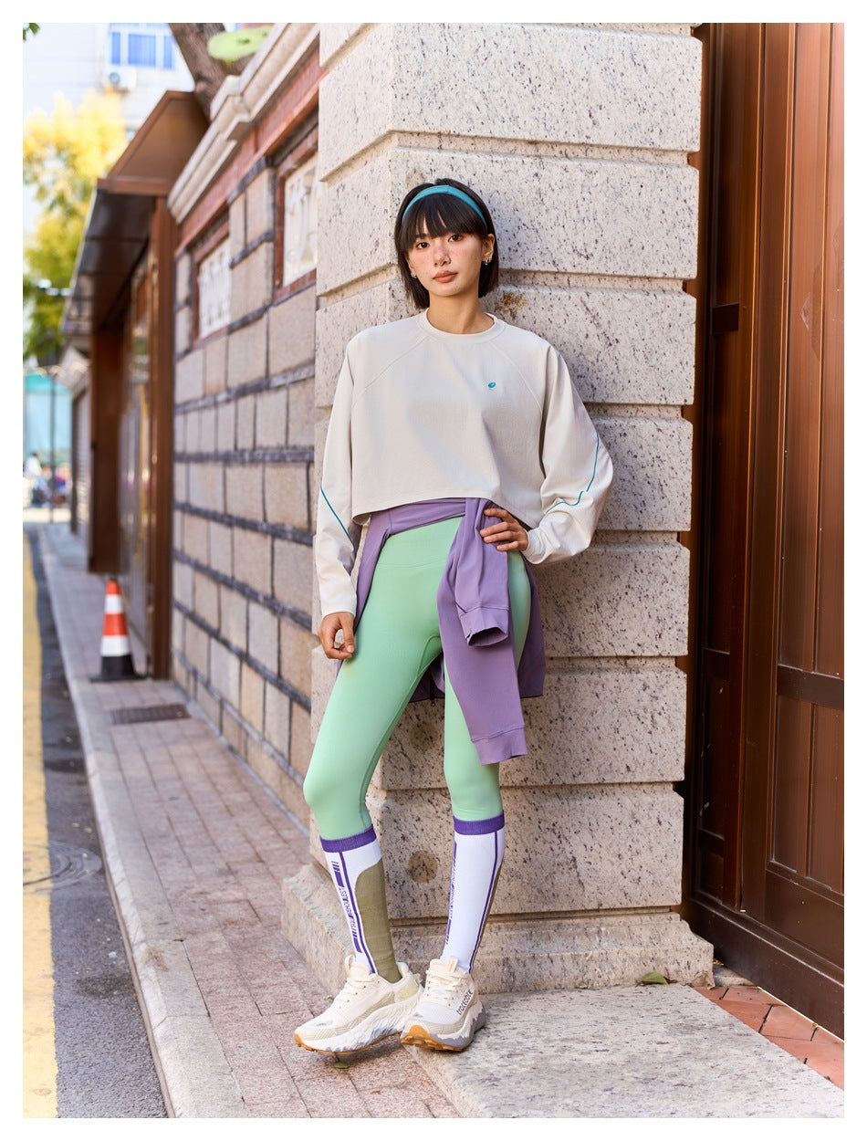 Woman wearing a cream cropped sweatshirt and mint leggings, with a lilac top tied around her waist.