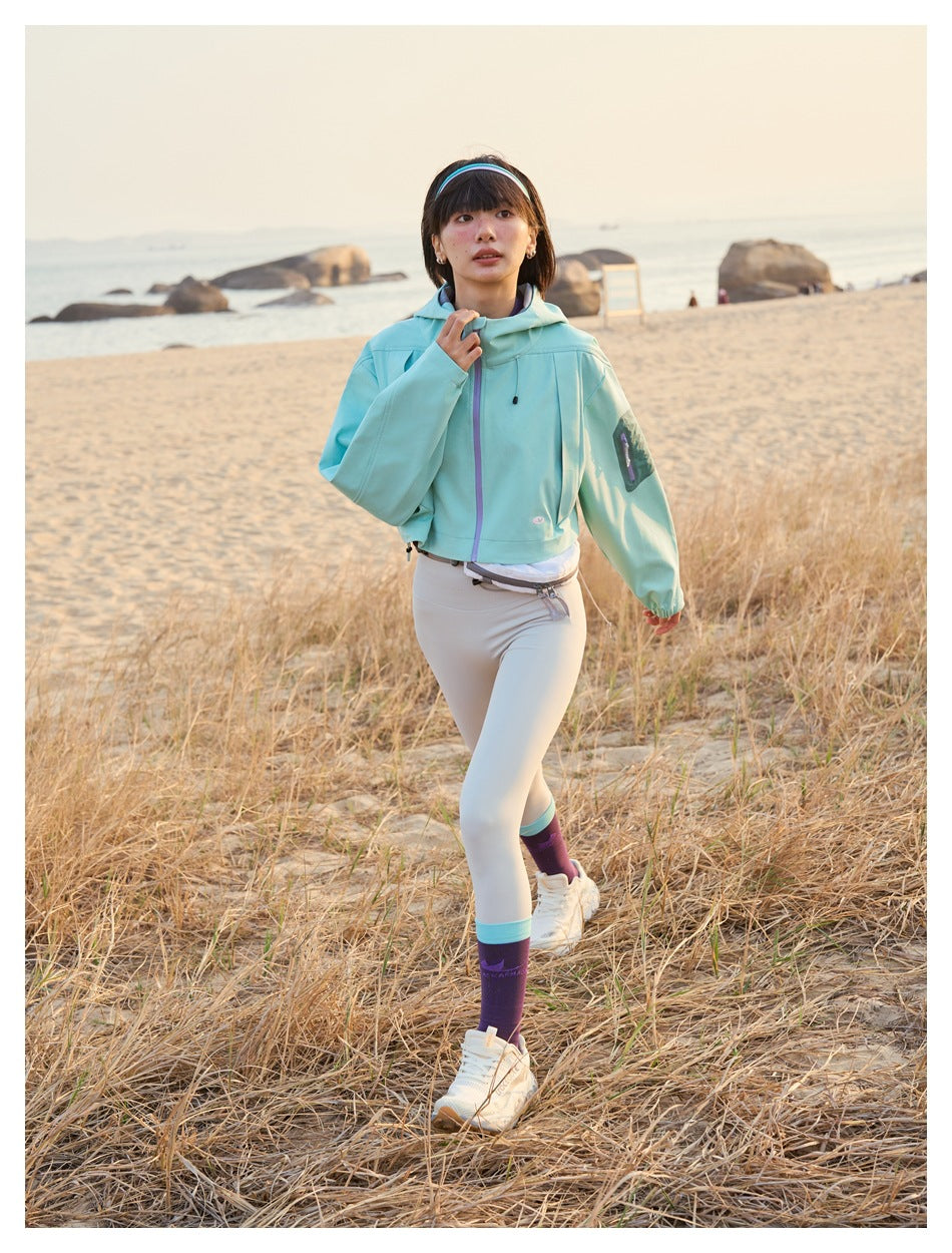 Woman walking on a beach in a cropped aqua hooded jacket, pale leggings, and purple socks.