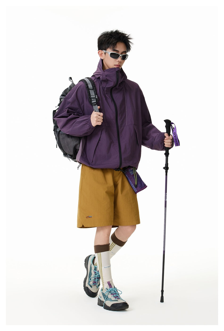 Model wearing a plum high-collar jacket and mustard shorts with a backpack and trekking pole.