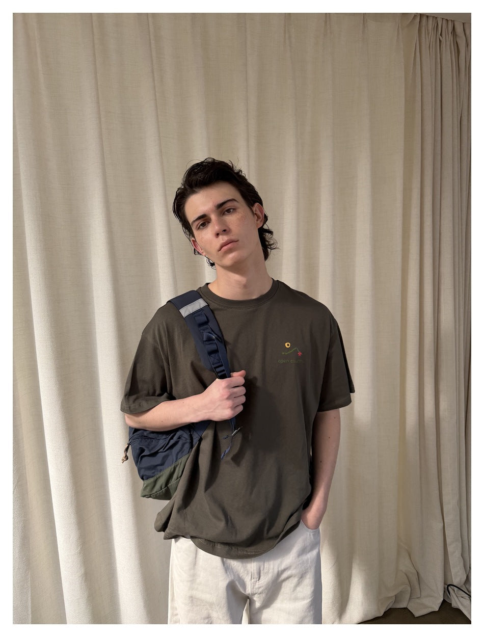 Close view of a model wearing a dark olive T-shirt with a small multicolor chest graphic.
