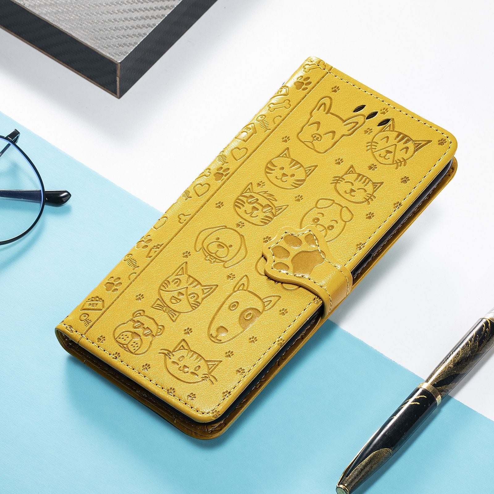 A yellow iPhone 16 Pro Max wallet case, featuring an embossed design of cute cats and dogs and a paw print clasp, rests on a blue and white surface alongside glasses and a pen.