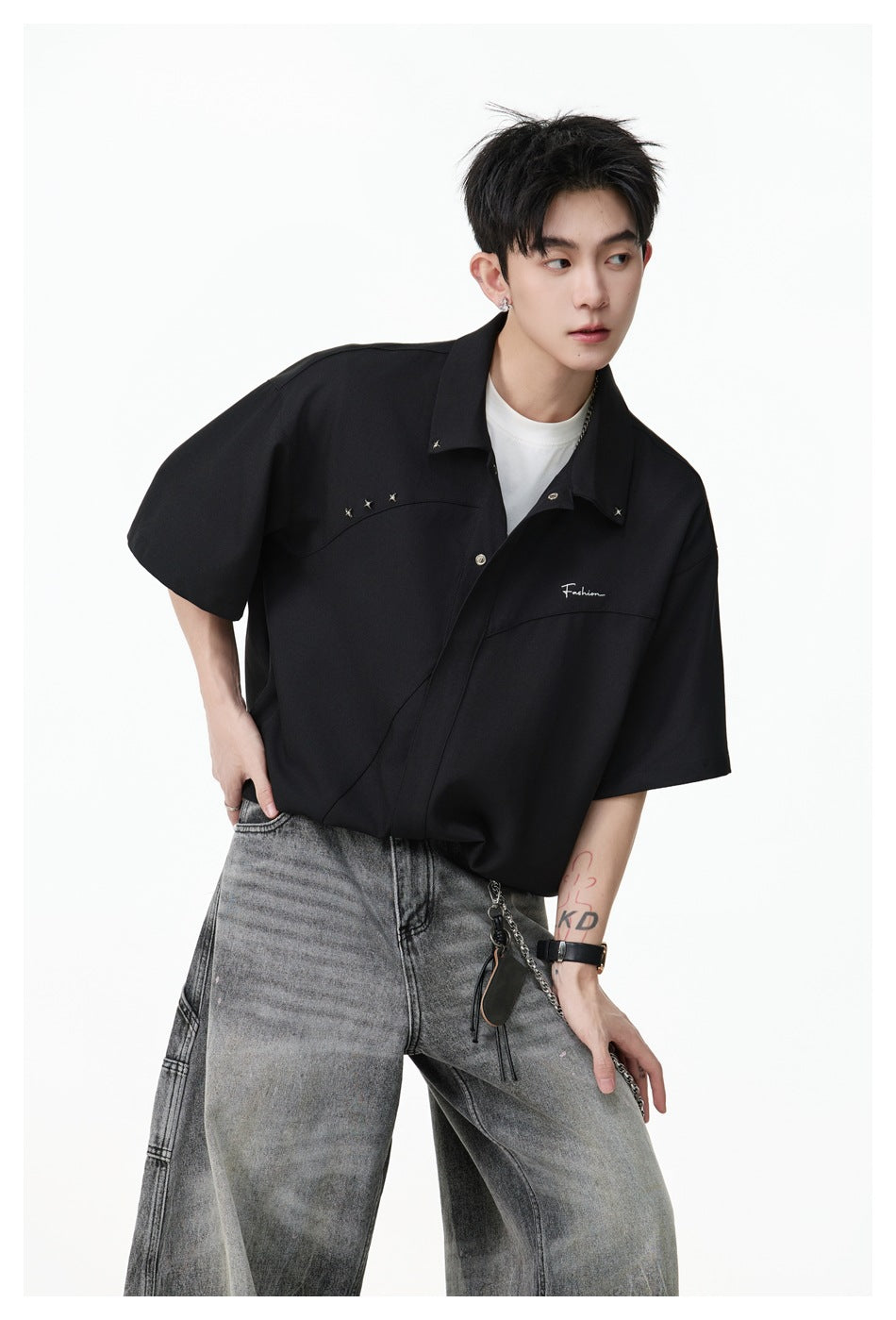 Model wearing the black short-sleeve shirt with star studs and chest embroidery over a white tee and gray wide-leg jeans