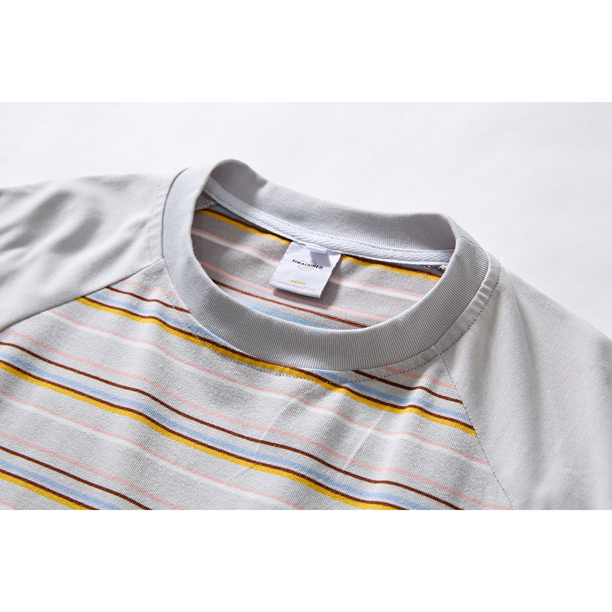 Close-up of a light gray raglan T-shirt collar and chest with thin yellow, blue, brown, and pink stripes.