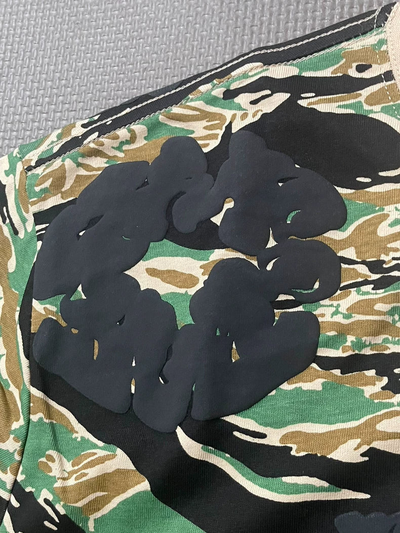 A close-up of the Denim Tears Green Camo All-Over Print T-Shirt, highlighting its textured gray background, green, black, and brown camouflage pattern with thick glossy black abstract shapes overlaying the bold design.