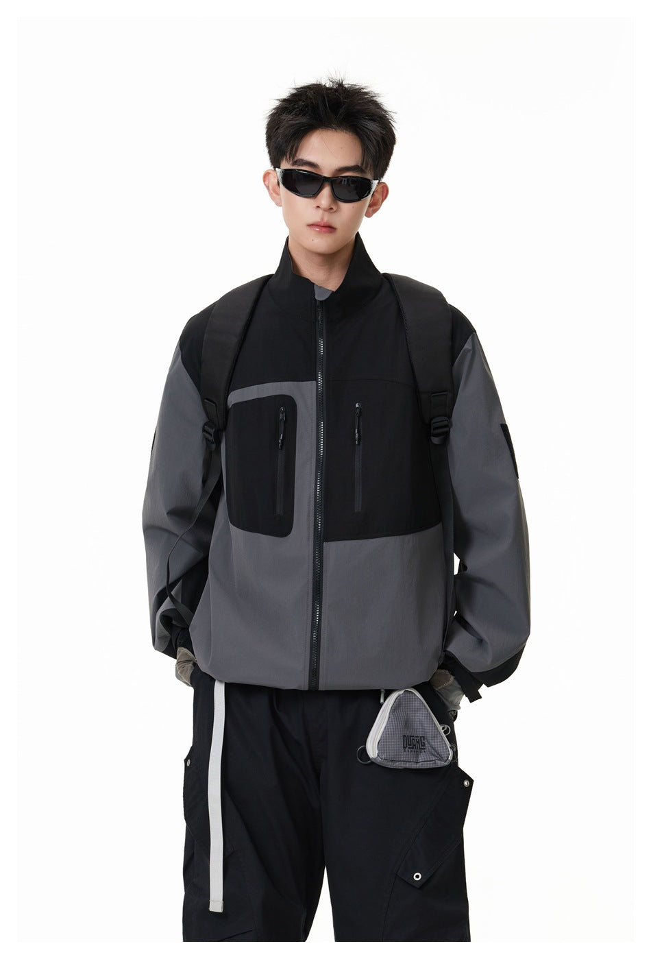 Model wearing a black and charcoal-gray color-block zip jacket with chest pockets and a high collar.