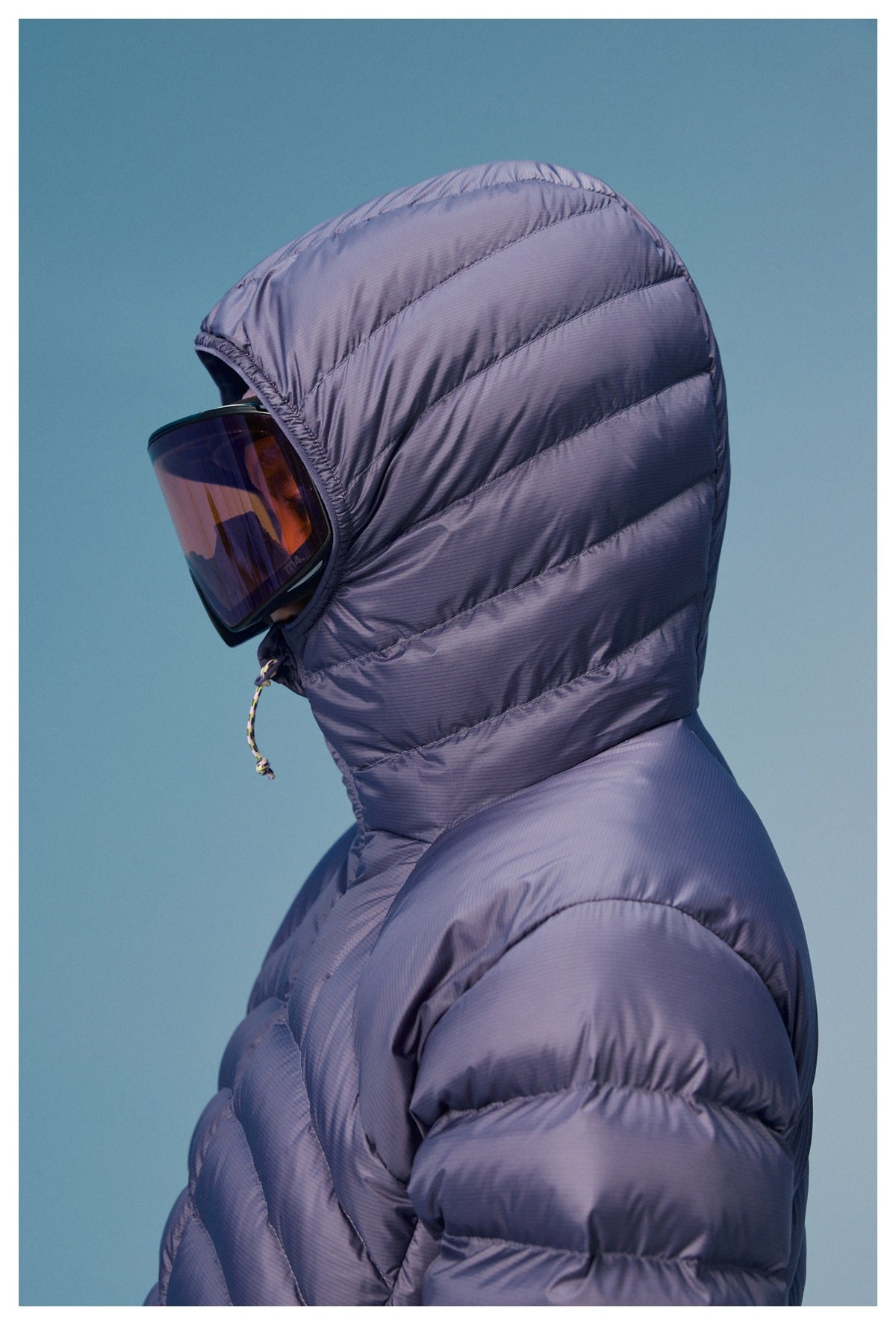 Side close-up of a person wearing a lavender quilted puffer jacket with a raised hood and ski goggles.