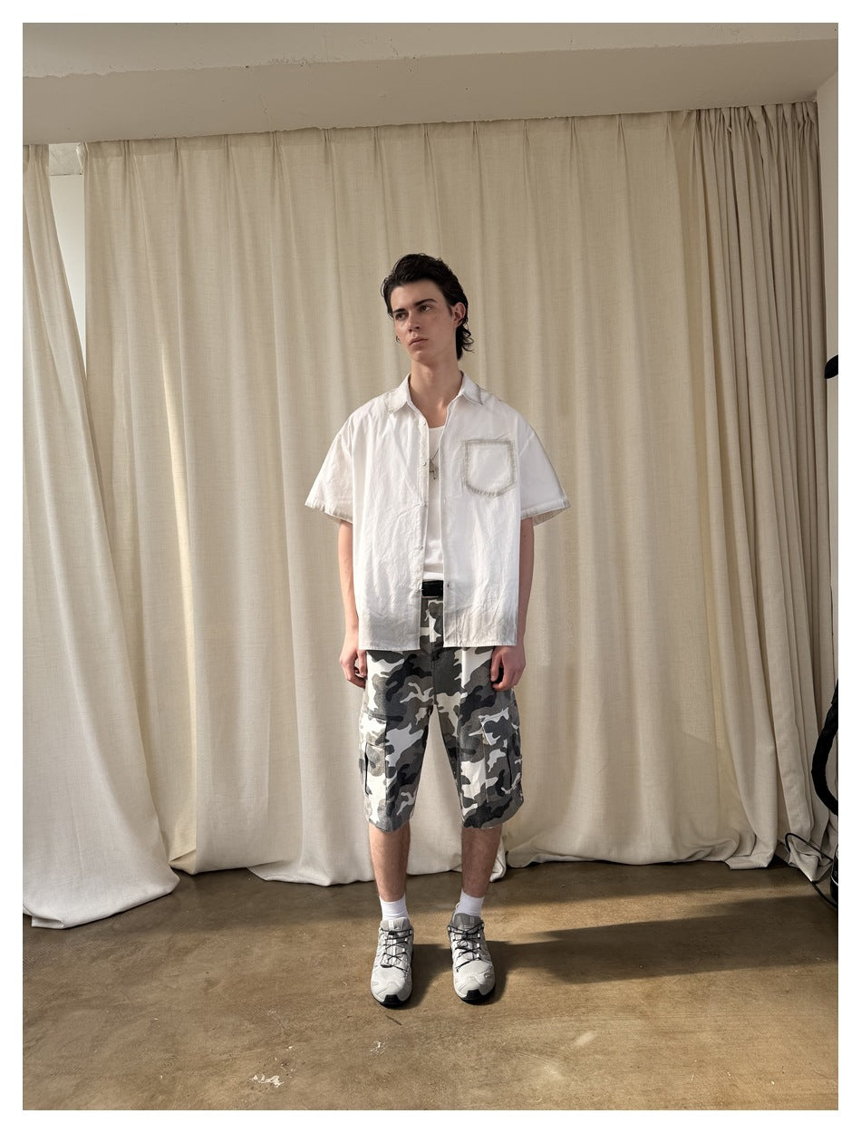 Man wearing an open white short-sleeve shirt with gray distressed edging over a white tee, camo shorts and sneakers