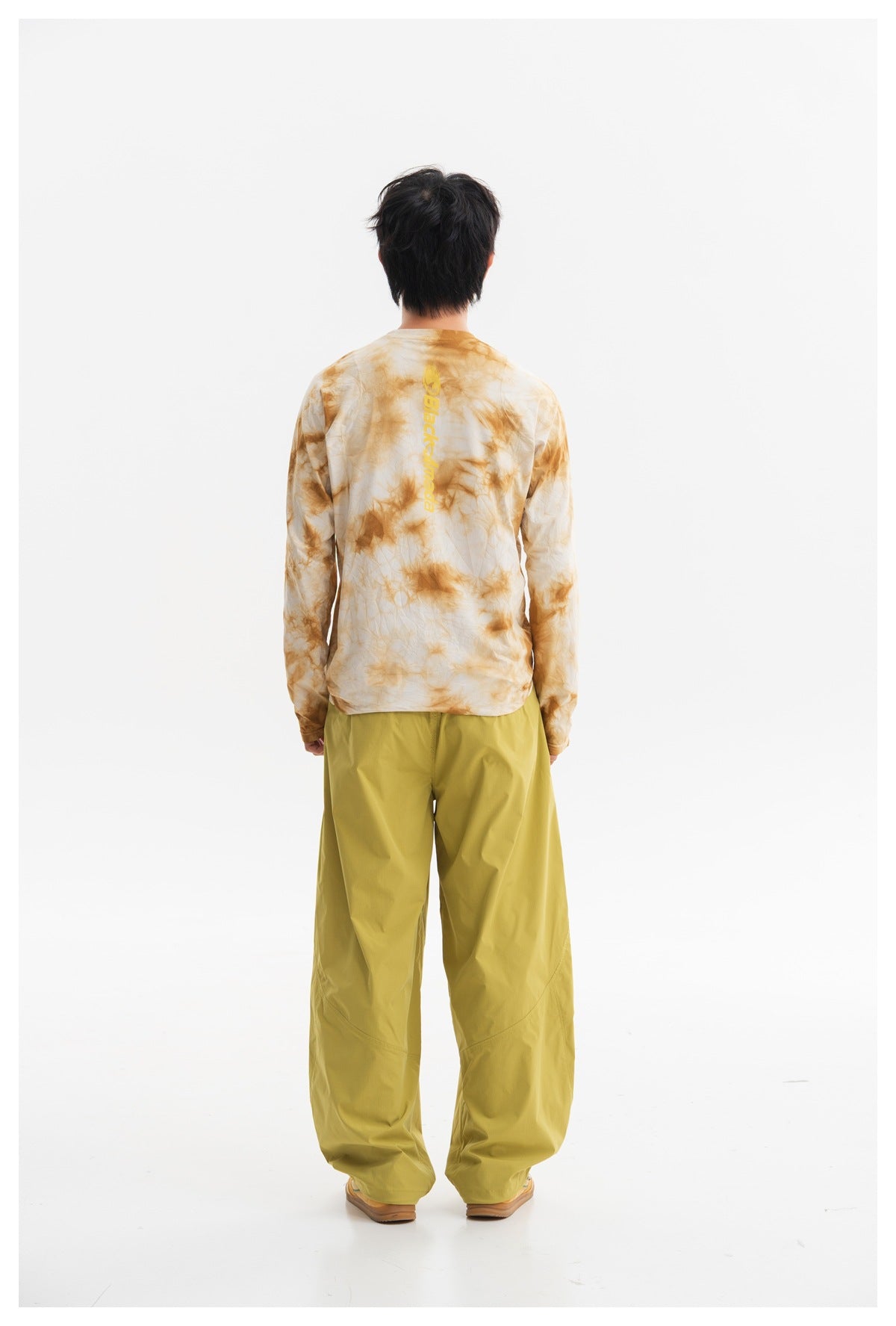 Back view of a model wearing a cream and ochre tie-dye long-sleeve shirt with a vertical yellow logo.