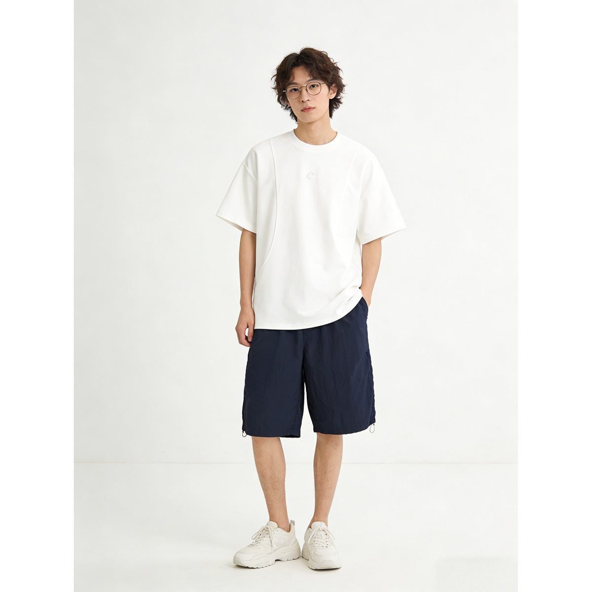 Model wearing an oversized white T-shirt with a small chest emblem and navy shorts, shown from the front.