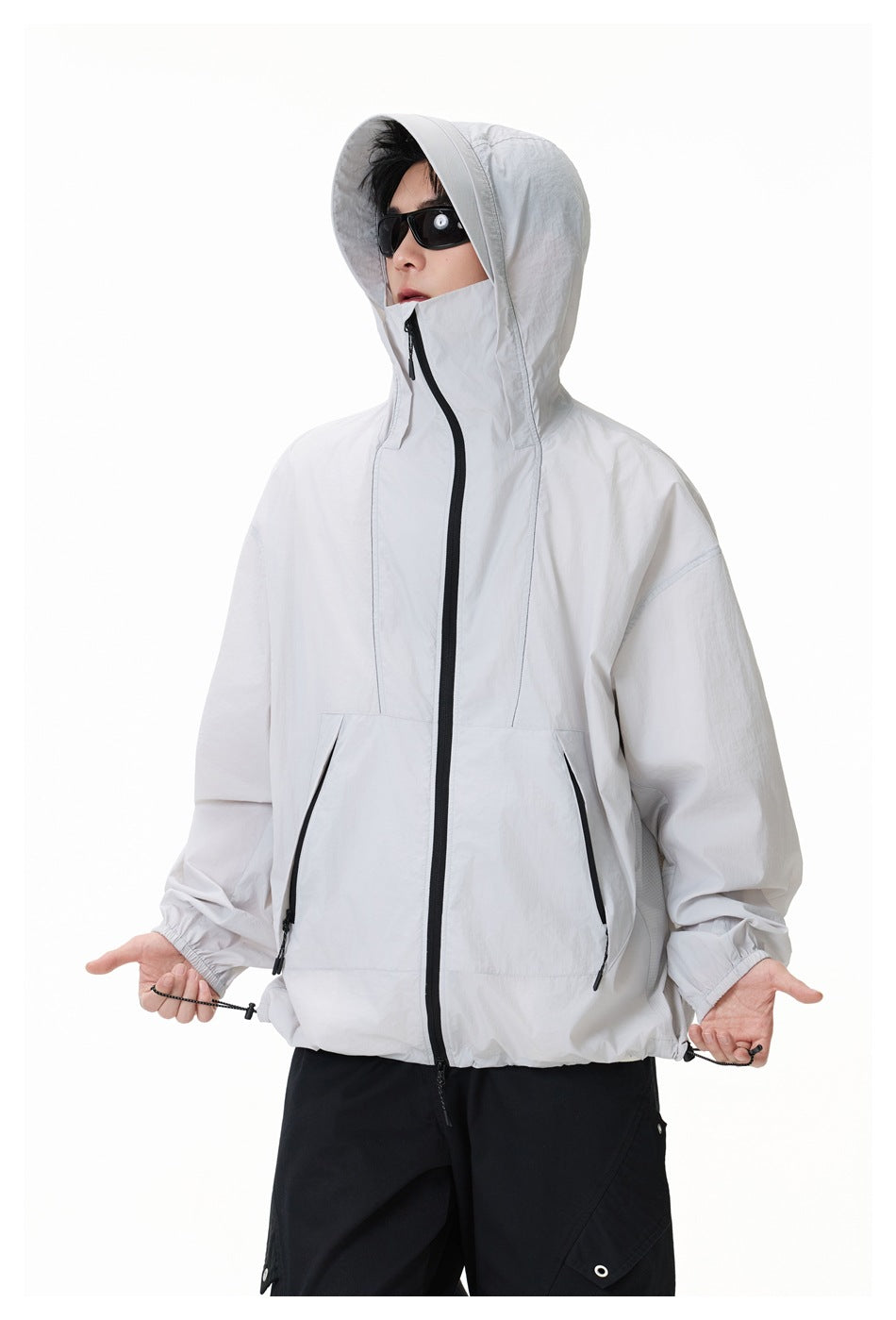 Model wearing a light gray hooded jacket with the hood raised, black sunglasses, and visible waist drawcords.