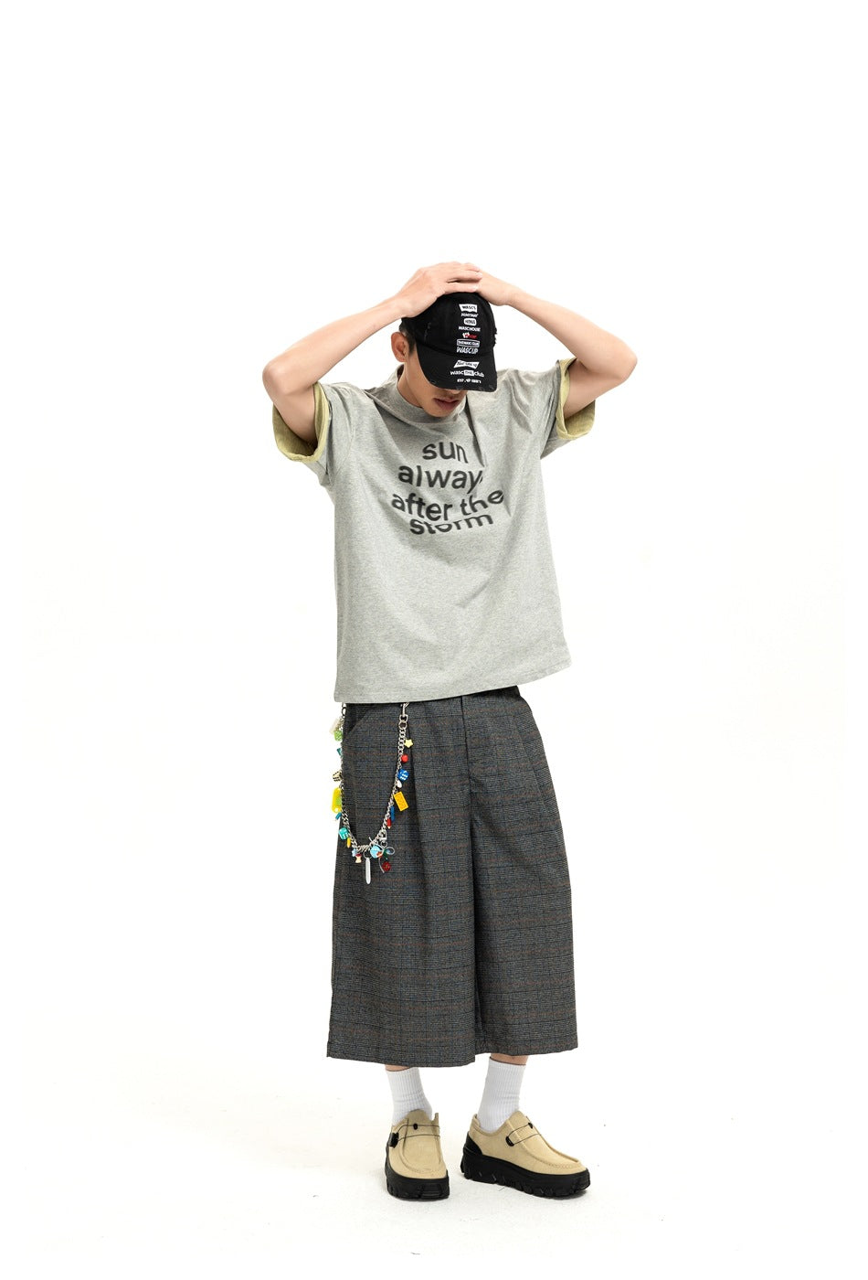 Model with hands on head wearing dark gray plaid wide-leg shorts, gray graphic T-shirt, cap and beige shoes