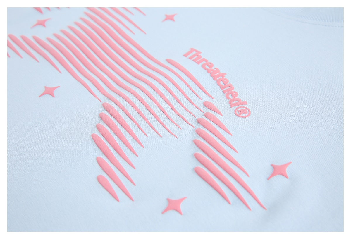 Angled close-up of the raised pink striped graphic, stars, and “Threatened®” lettering on a pale blue T-shirt.