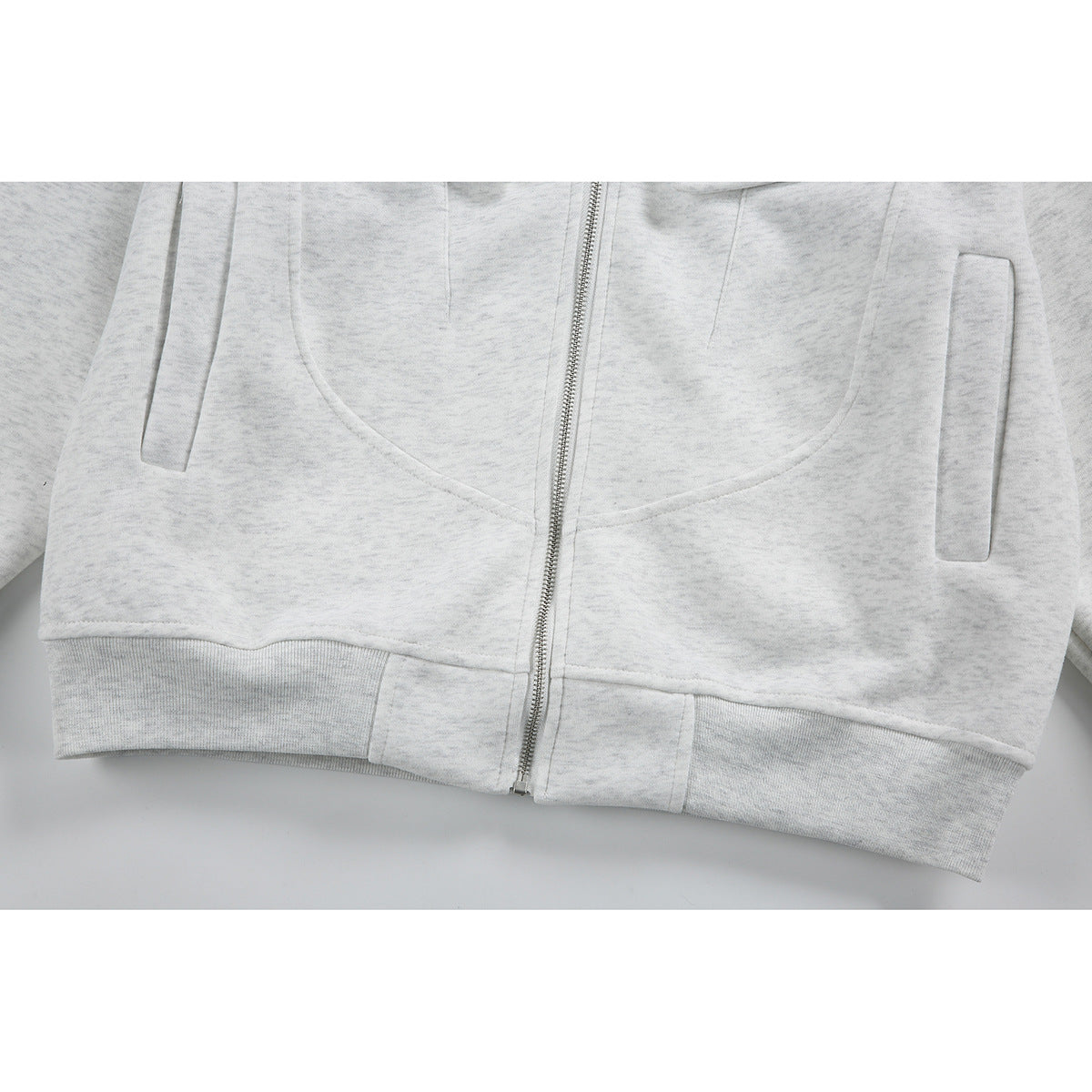 Close-up of the light gray jacket's lower front, showing the zipper, curved seams, vertical pockets, and ribbed hem.