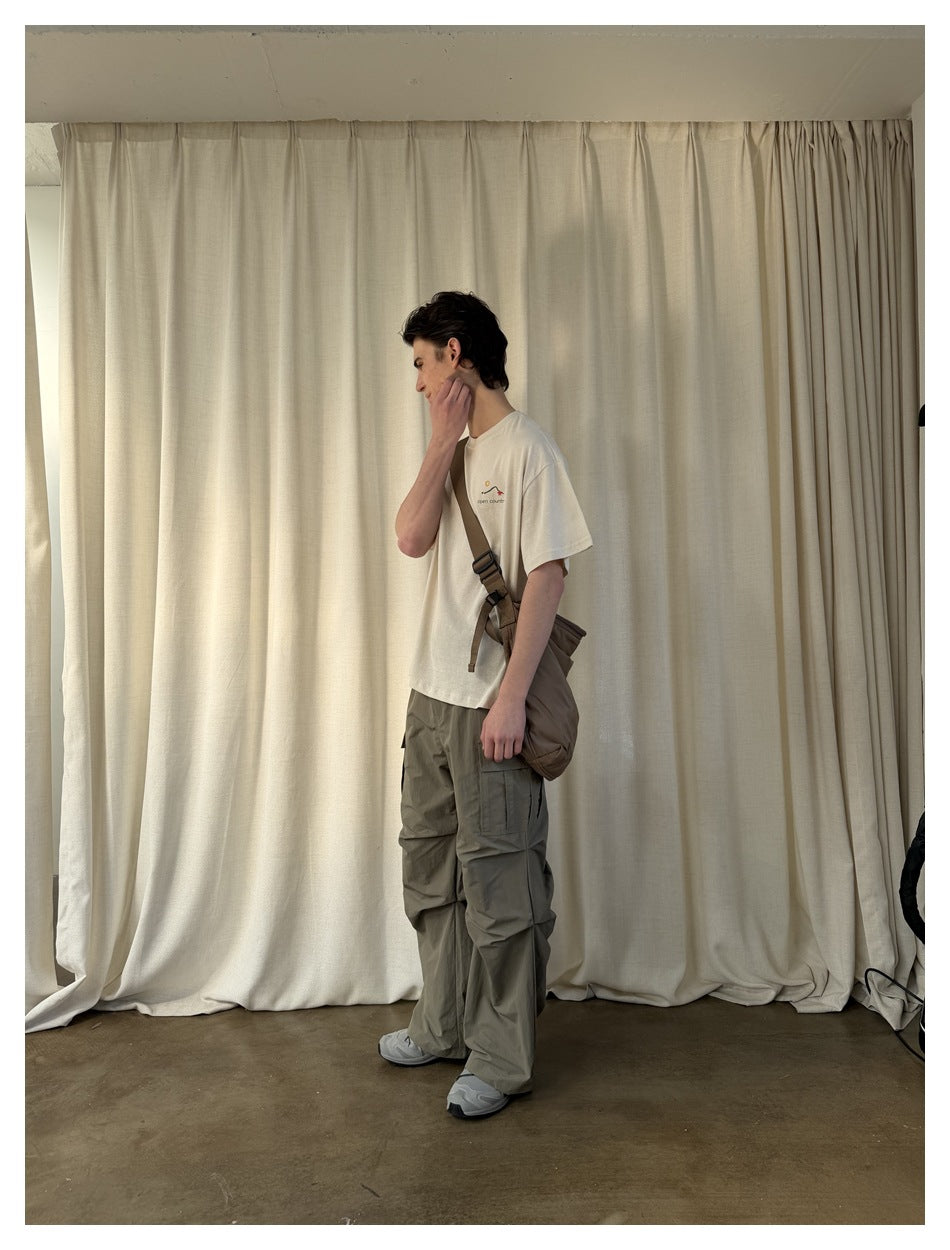 Side view of a model in a cream graphic T-shirt, gray-olive cargo pants, and a brown crossbody bag.