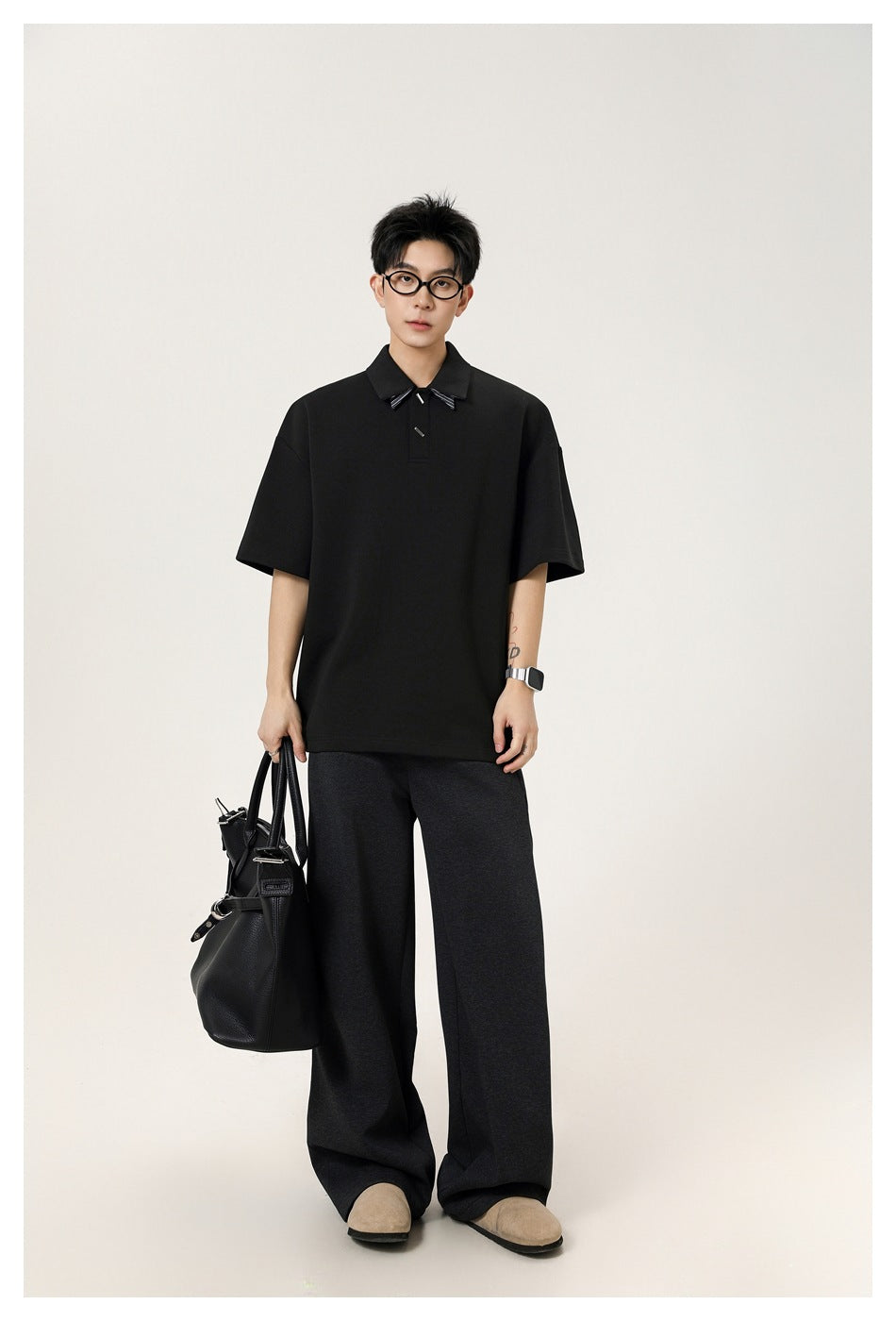 Model wearing a black short-sleeve polo with striped collar detail, wide dark gray trousers, and holding a black tote bag