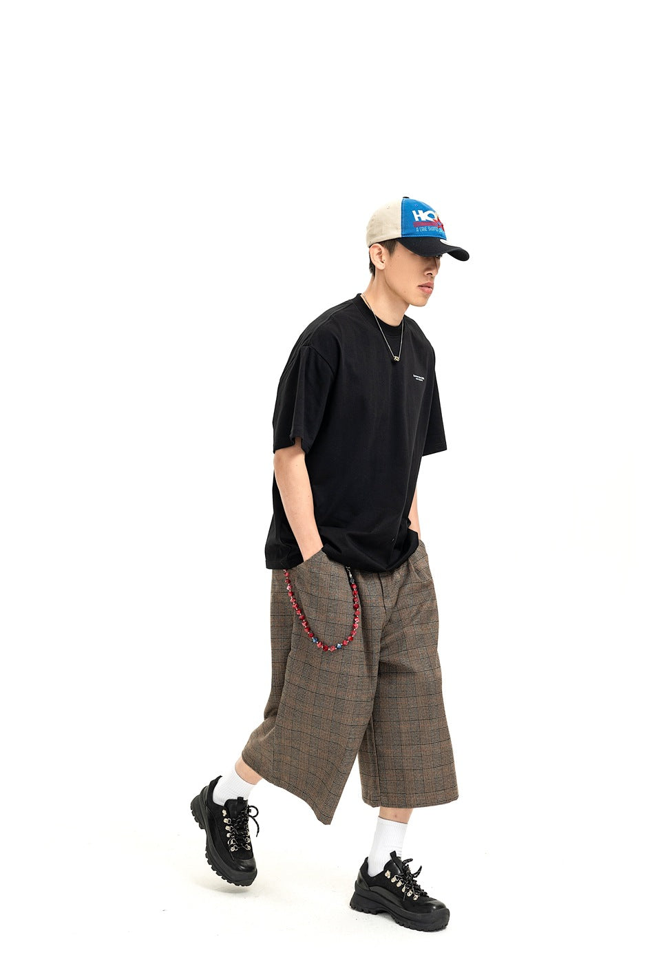 Model in a black T-shirt and brown plaid wide-leg shorts, shown walking in a color-block cap.