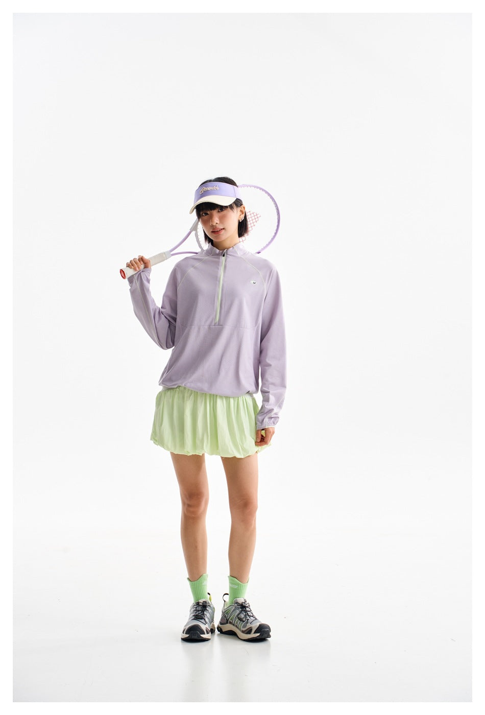 Full-length model in a lavender half-zip top and pale green bubble skirt, carrying a tennis racket over one shoulder.