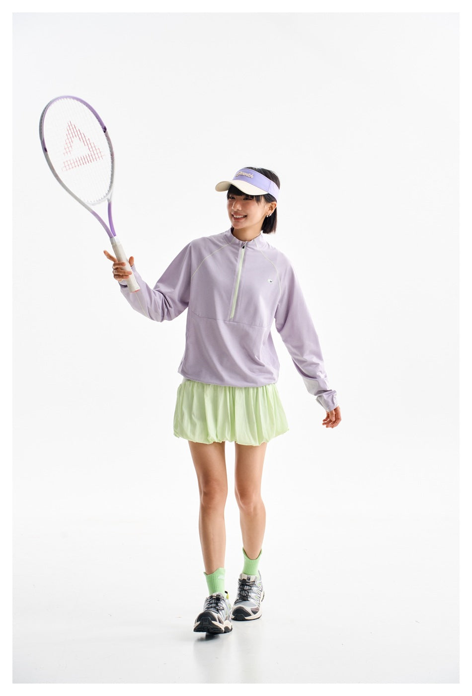 Model in a lavender half-zip top and pale green bubble skirt, holding a tennis racket.