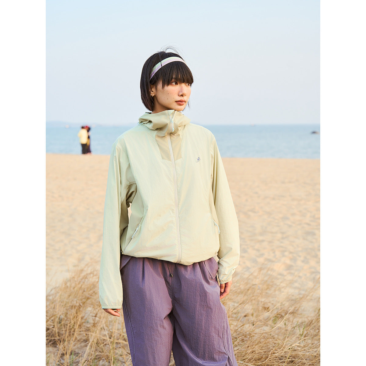 Model wearing a pale yellow hooded zip jacket with purple trousers on a beach.