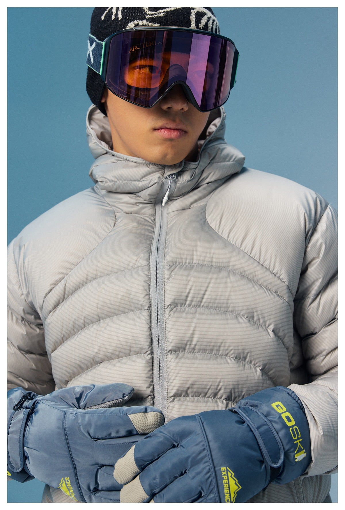 Close-up of a model wearing a light gray quilted puffer jacket with a high collar and blue ski gloves.