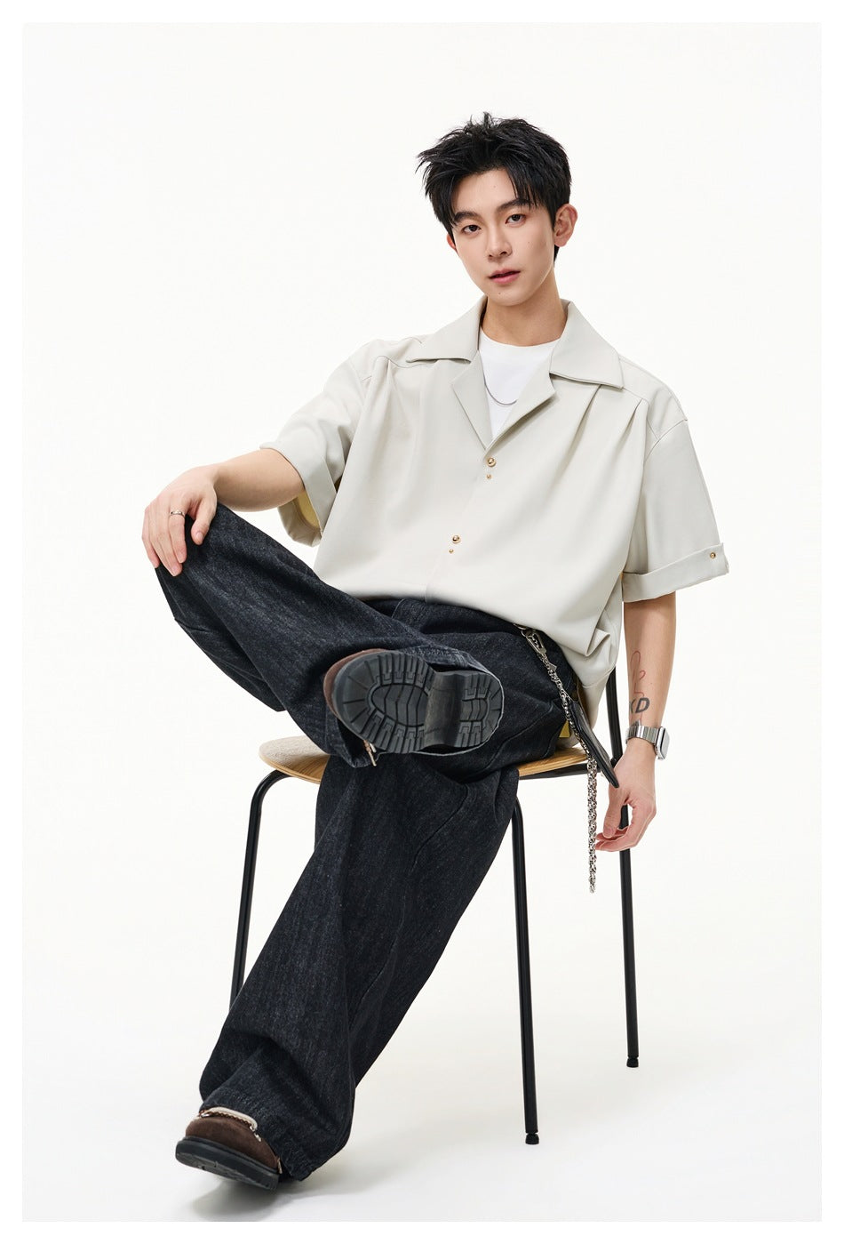 Seated model wearing a light cream oversized short-sleeve shirt with wide lapels and gold-tone buttons.