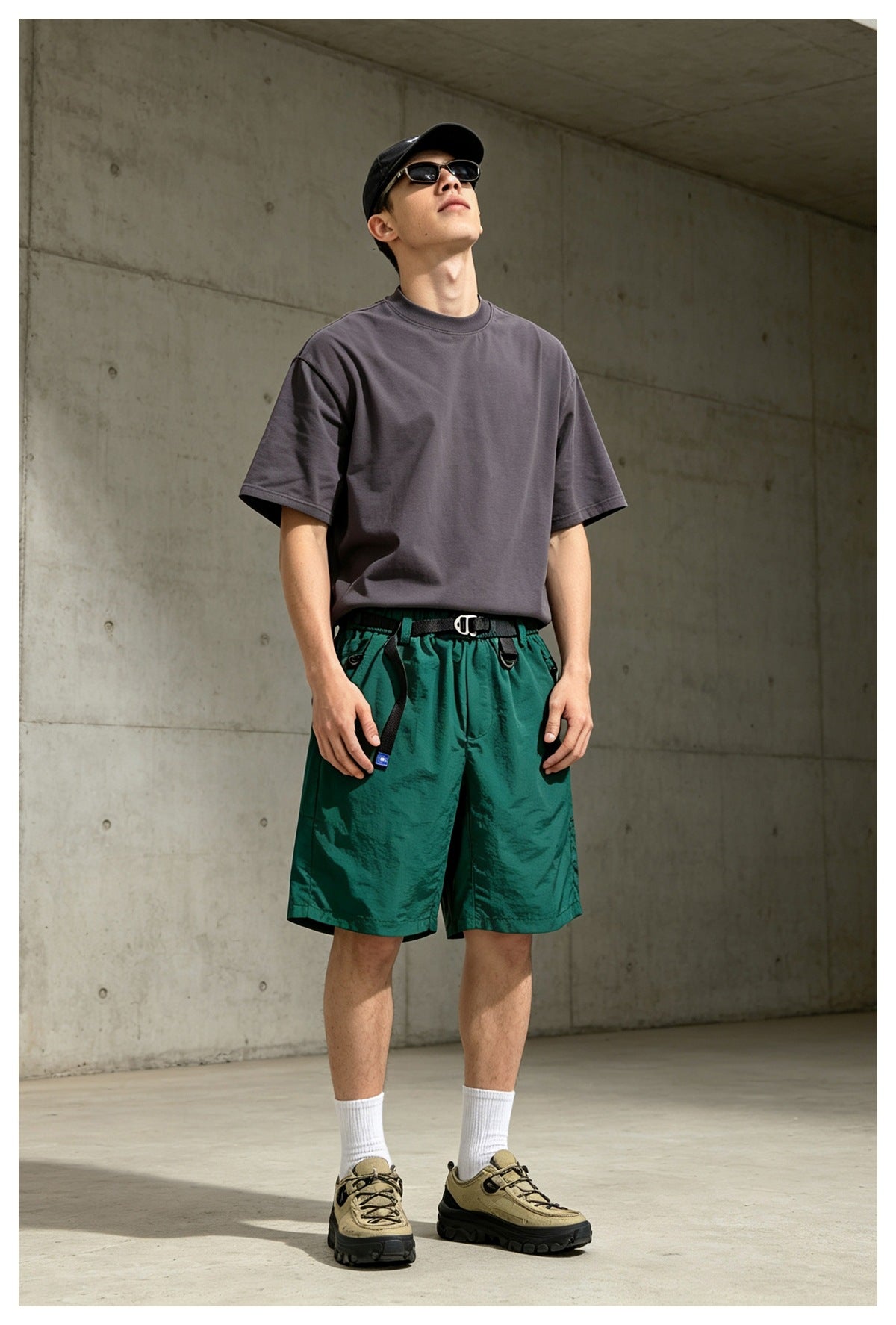 Model wearing green belted shorts with a gray t-shirt, cap, sunglasses, white socks and sneakers against a concrete wall