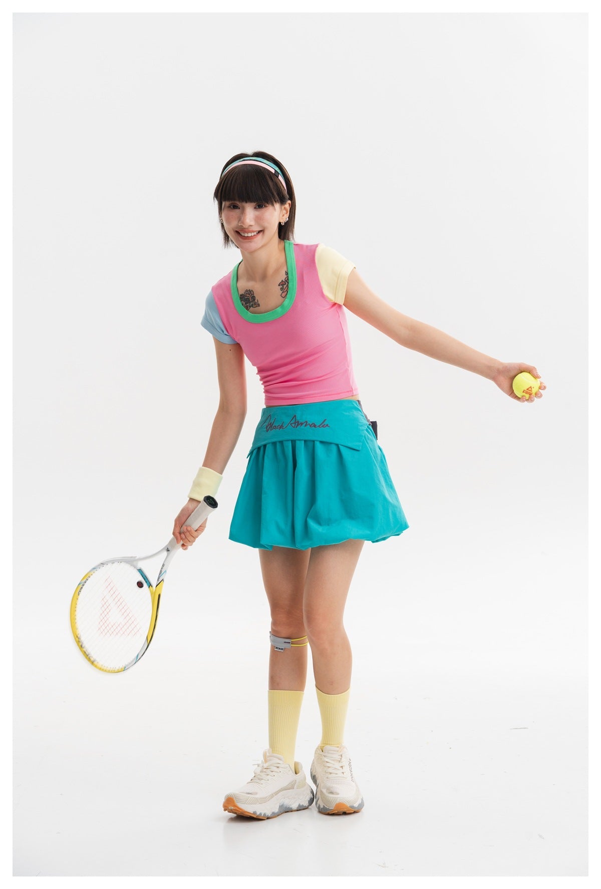 Model wearing a pink top with green neckline and contrasting sleeves, paired with a turquoise skirt, holding a tennis racket