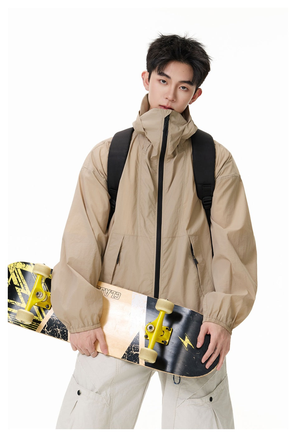 Model wearing a beige hooded jacket with a high zipped collar and black backpack, holding a skateboard.