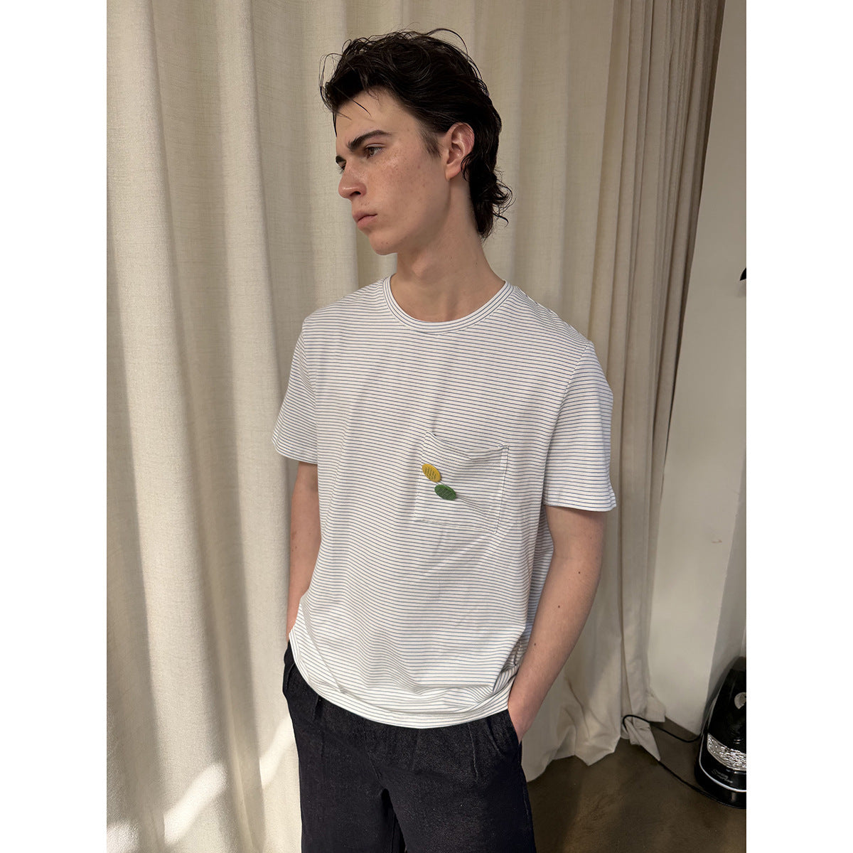 Model wearing a white T-shirt with thin blue stripes, a chest pocket and small yellow and green decorations.