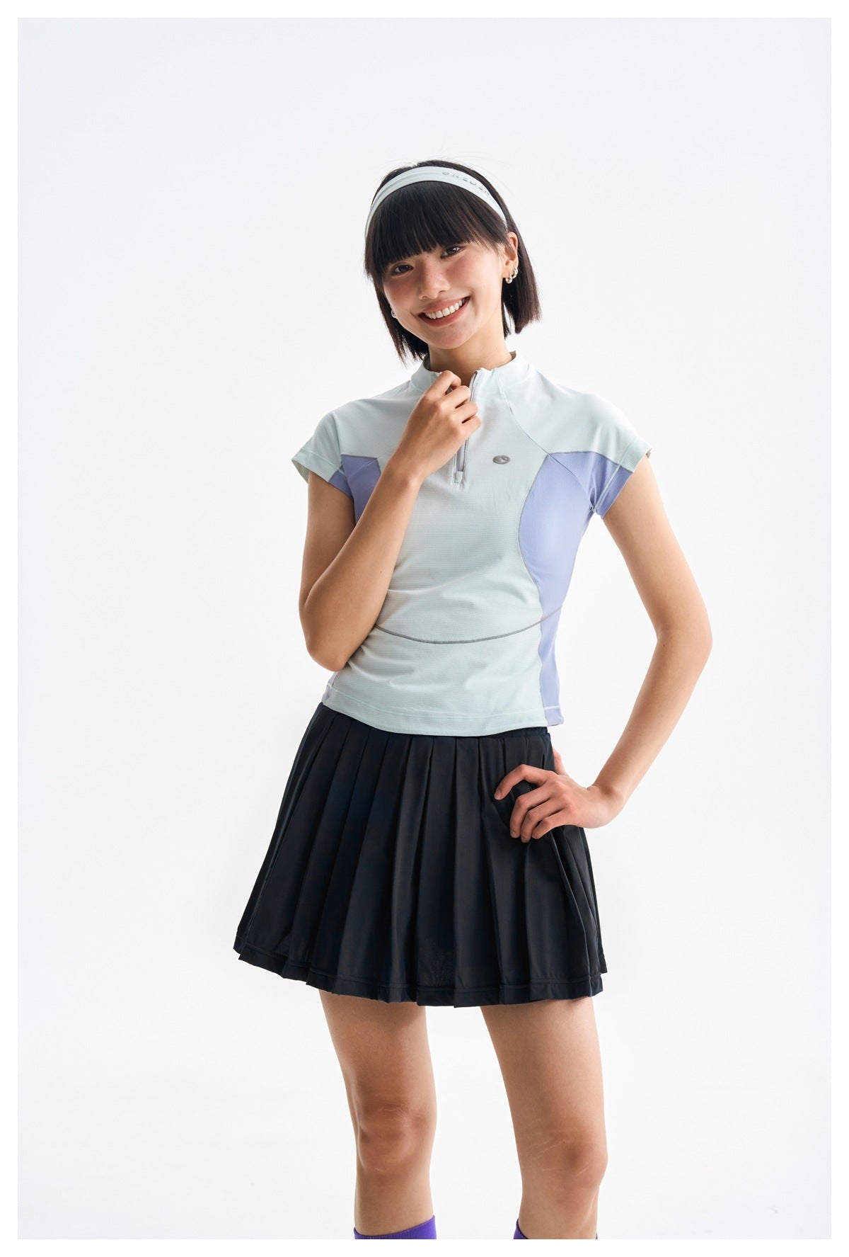 Model wearing a pale blue zip-neck top with lavender side panels and a black pleated skirt.