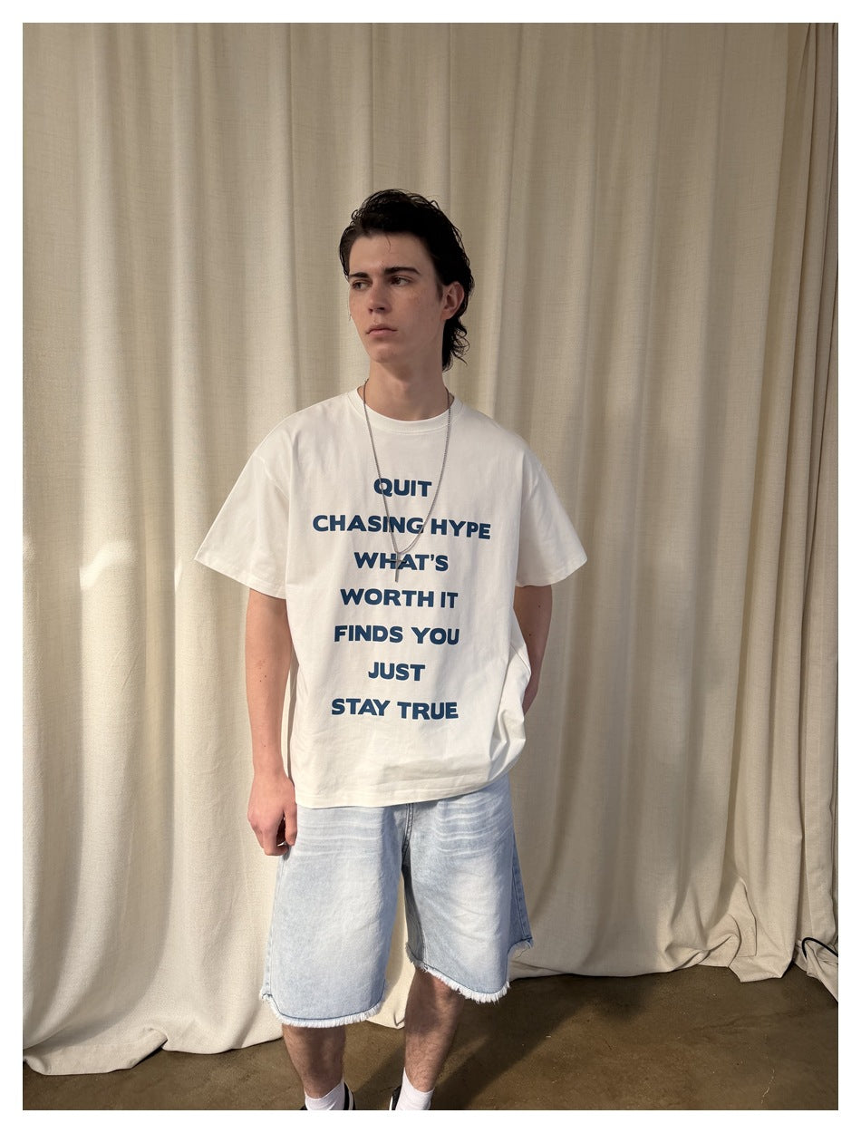 Model wearing a white oversized T-shirt with bold navy text, pale denim shorts, and a silver necklace.