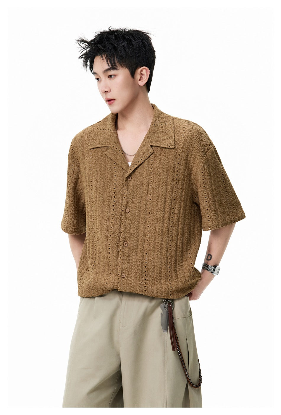 Model wearing a brown short-sleeve eyelet shirt with beige trousers, shown from a three-quarter angle.