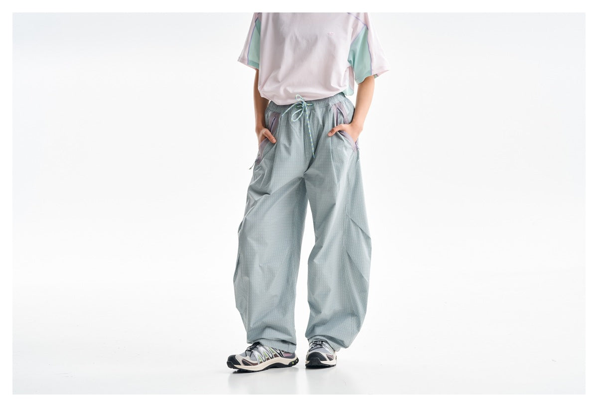 Front view of a model wearing loose pale blue-gray check trousers with a drawstring waist and contrast zip pockets.