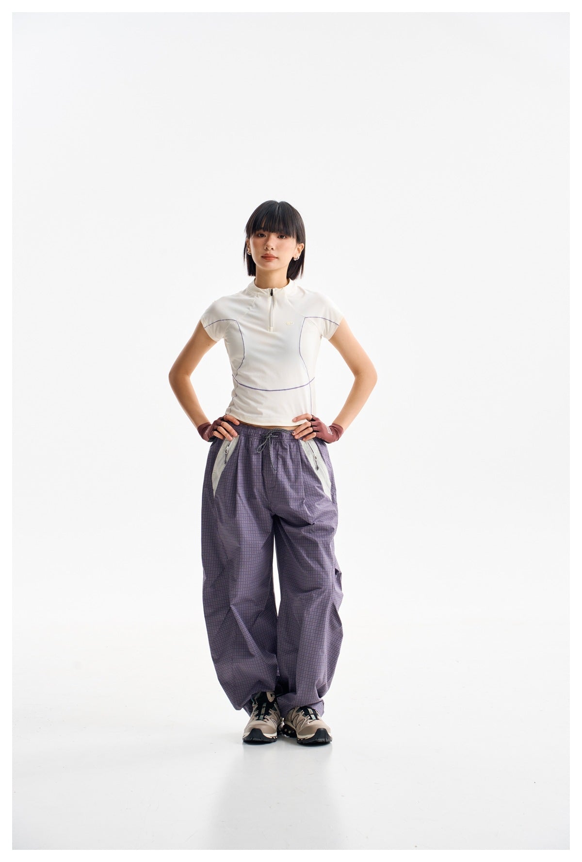 Full-length front view of a model wearing loose purple check trousers with light contrast pocket panels and a white zip-neck