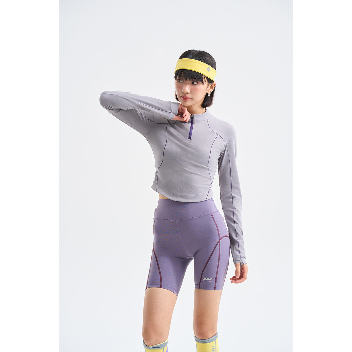Model wearing a gray-lilac quarter-zip top and purple fitted shorts with dark contrast seams.
