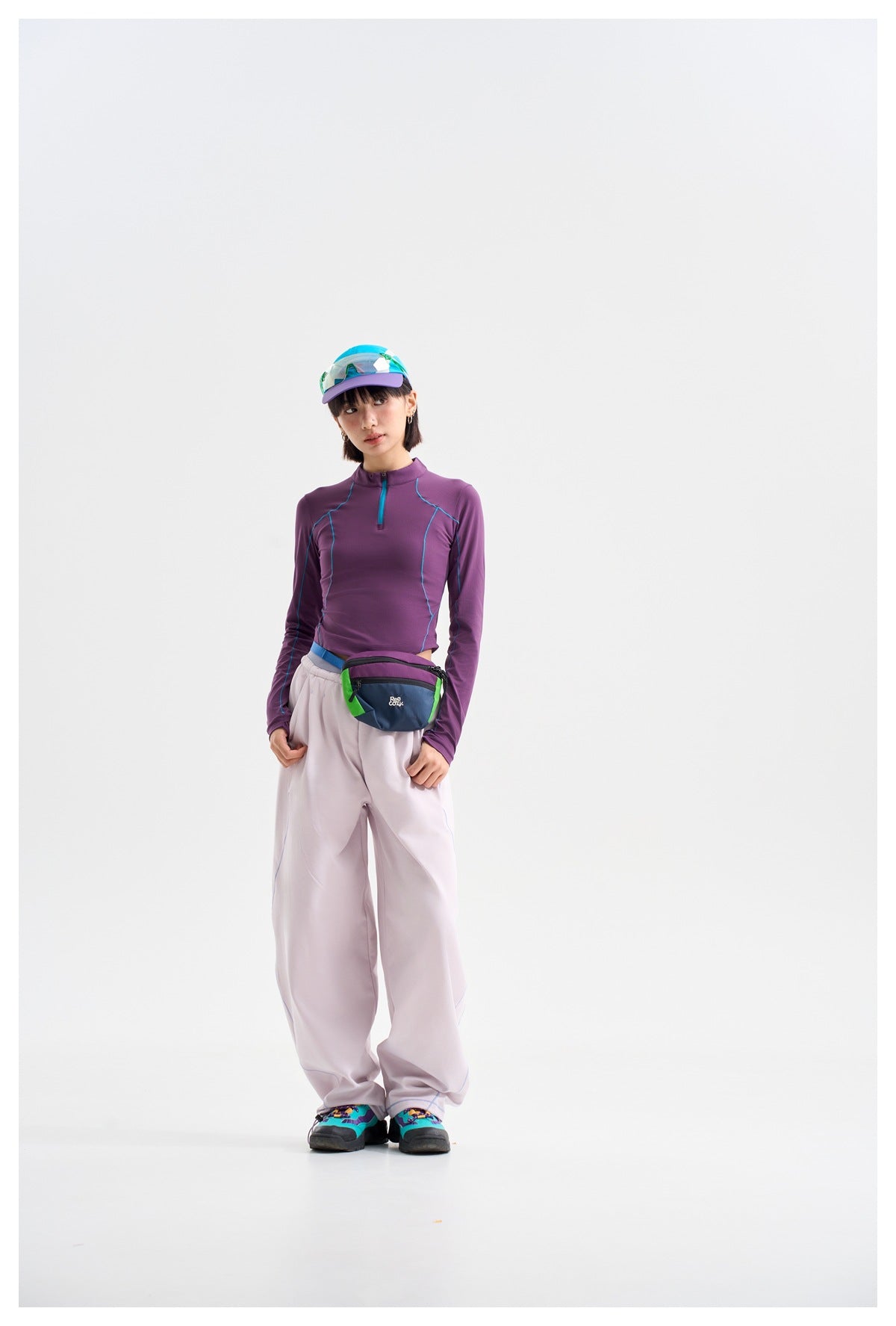 Model wearing a purple long-sleeve quarter-zip top with teal seams and loose pale trousers, with a colorful waist bag.