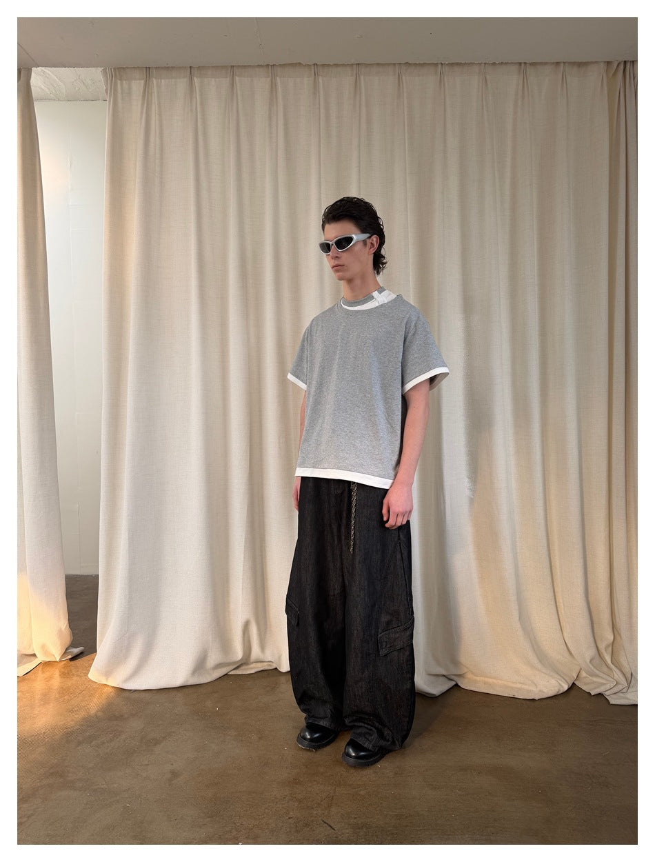 Model in a gray T-shirt with white trim and black cargo jeans, angled three-quarter view in front of a curtain