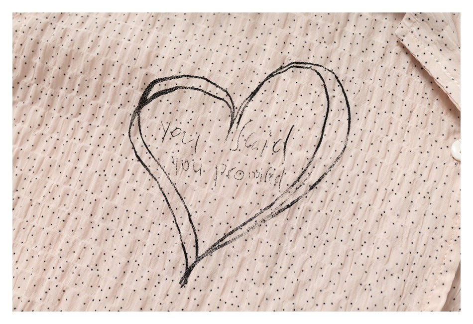 Close-up of a black outlined heart with faint lettering printed on the pale blush shirt’s dotted fabric.