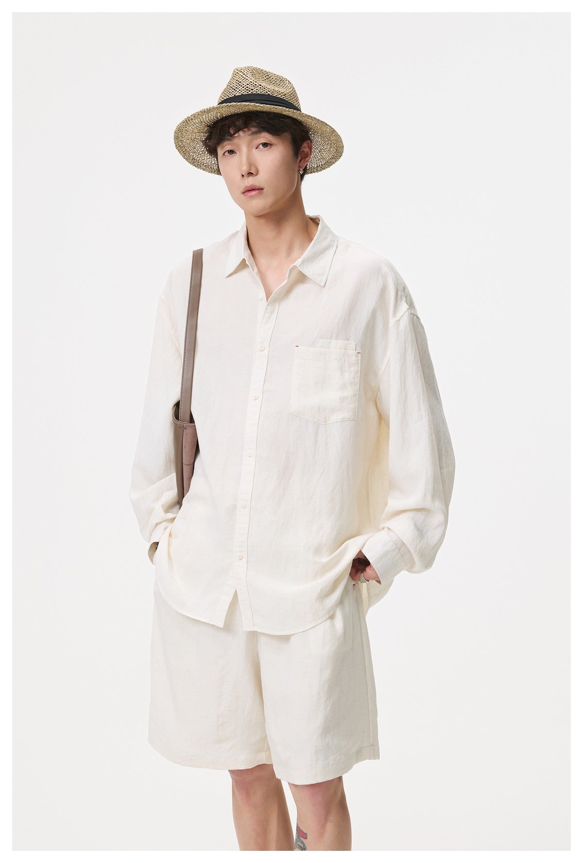 Man standing in beige shorts and a matching long-sleeve shirt with a straw hat and a shoulder bag