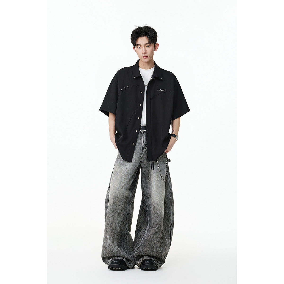 Model wearing a black short-sleeve button-up shirt with silver accents and oversized gray jeans, front view.