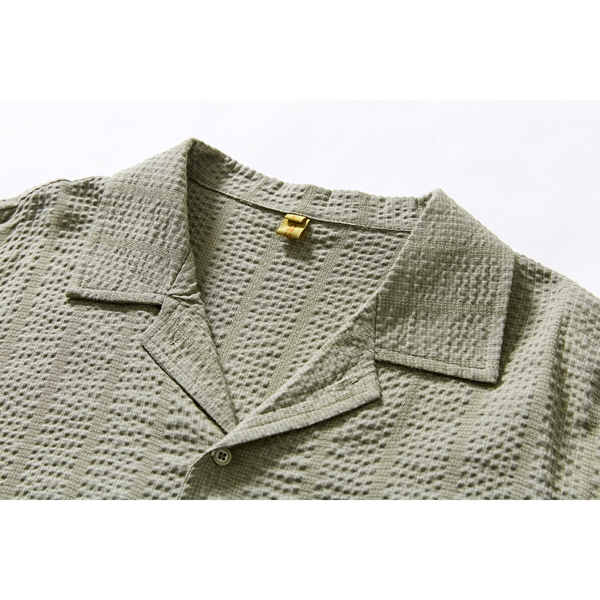 Close-up of a sage-gray textured short-sleeve shirt showing its notched collar, placket button, and small neck label.