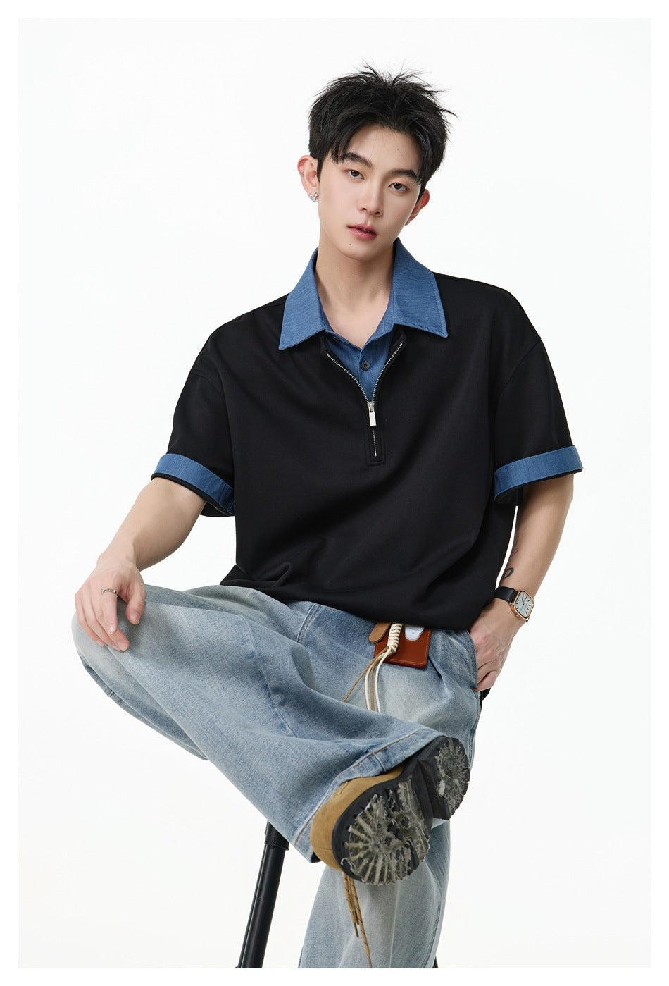 Seated model wearing a black short-sleeve quarter-zip polo with blue collar and cuffs, paired with light-wash jeans.