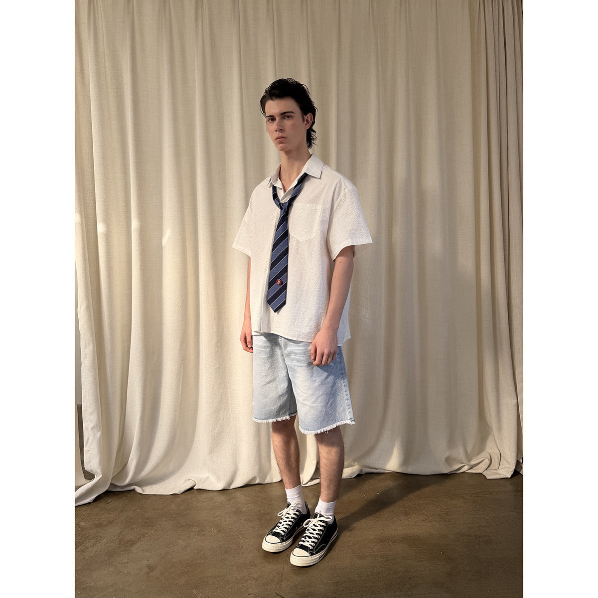 Full-length view of a model wearing a white short-sleeve shirt, navy striped tie, and light denim shorts.