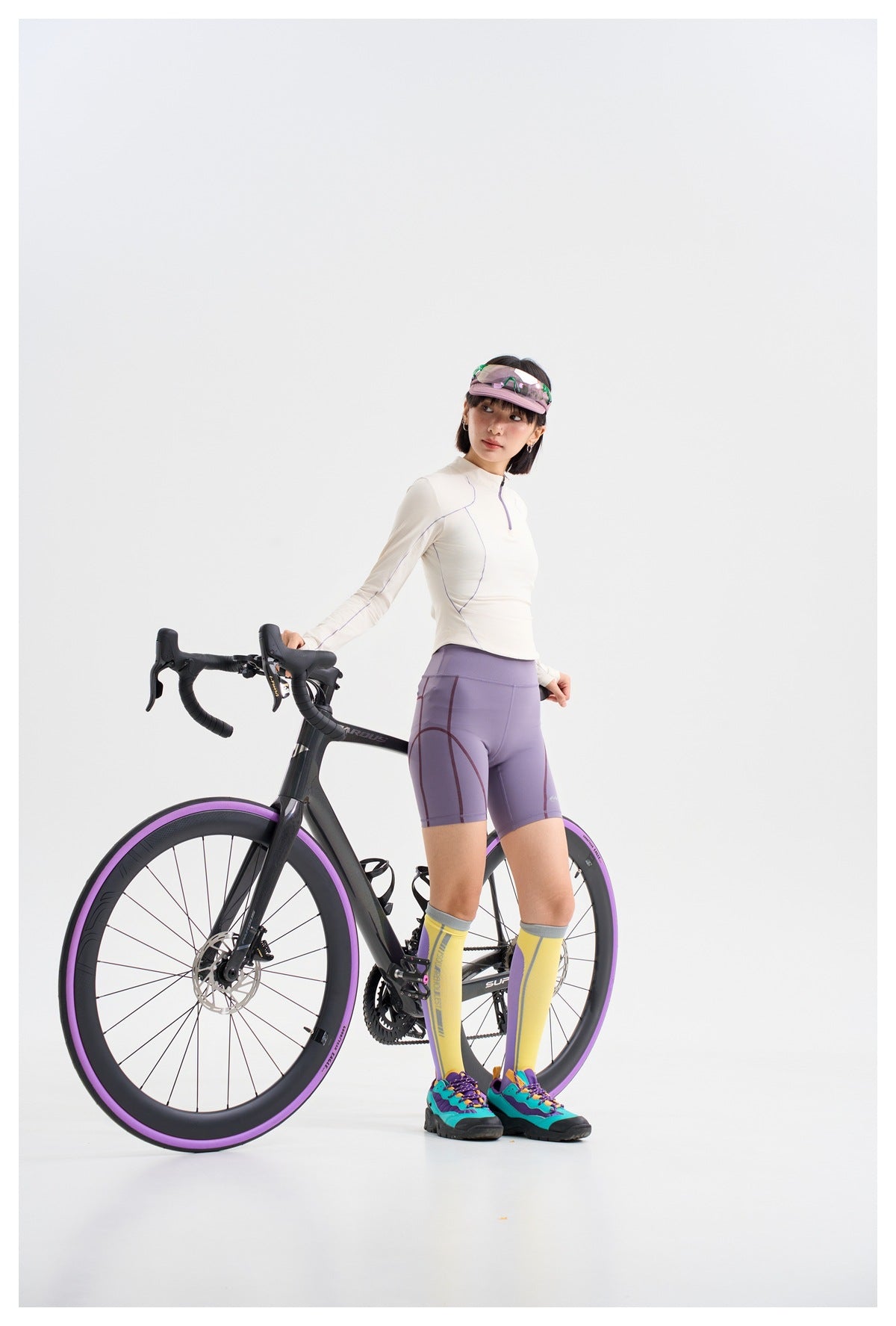 Model in a white long-sleeve top, lavender cycling shorts, and yellow socks, posing beside a black bicycle.