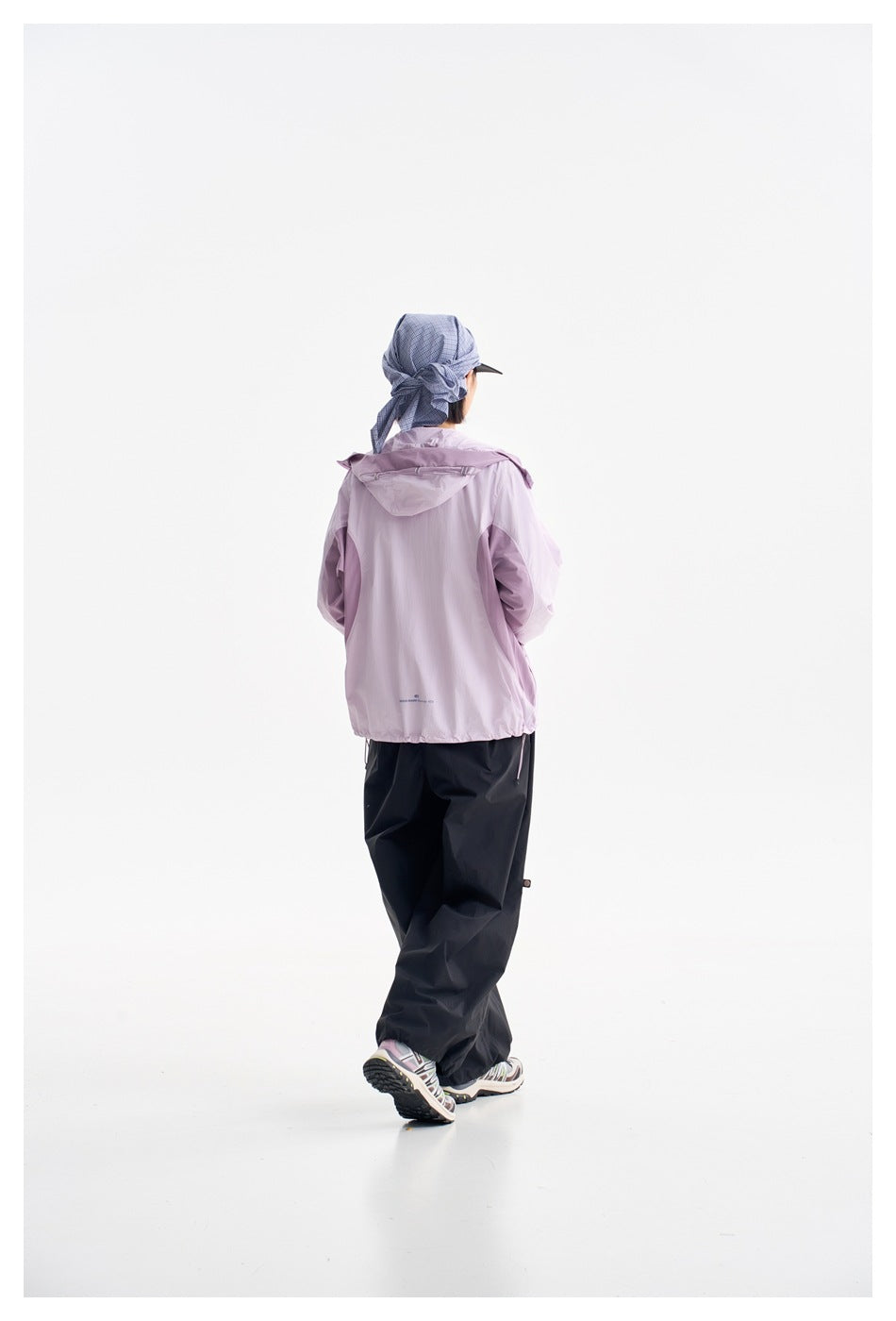 Rear view of a model wearing the lilac hooded jacket and loose black pants with a blue plaid cap.
